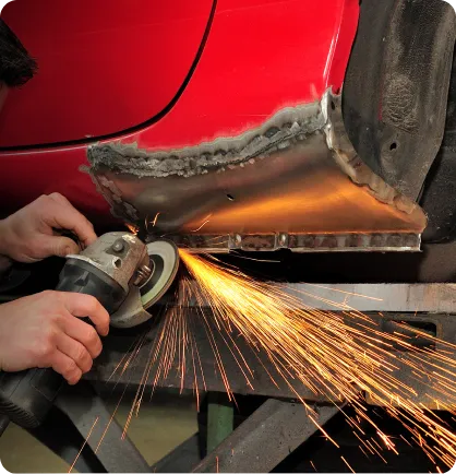 Prestador de serviço com uma lixadeira elétrica realizando o serviço automotivo de funilaria, em um carro vermelho, próximo ao assoalho do veículo.