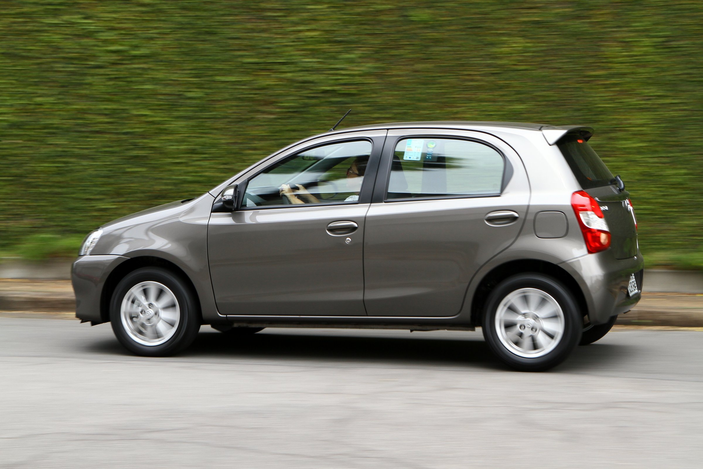 Toyota Etios X marrom de perfil em movimento na rua com um muro de plantas ao fundo