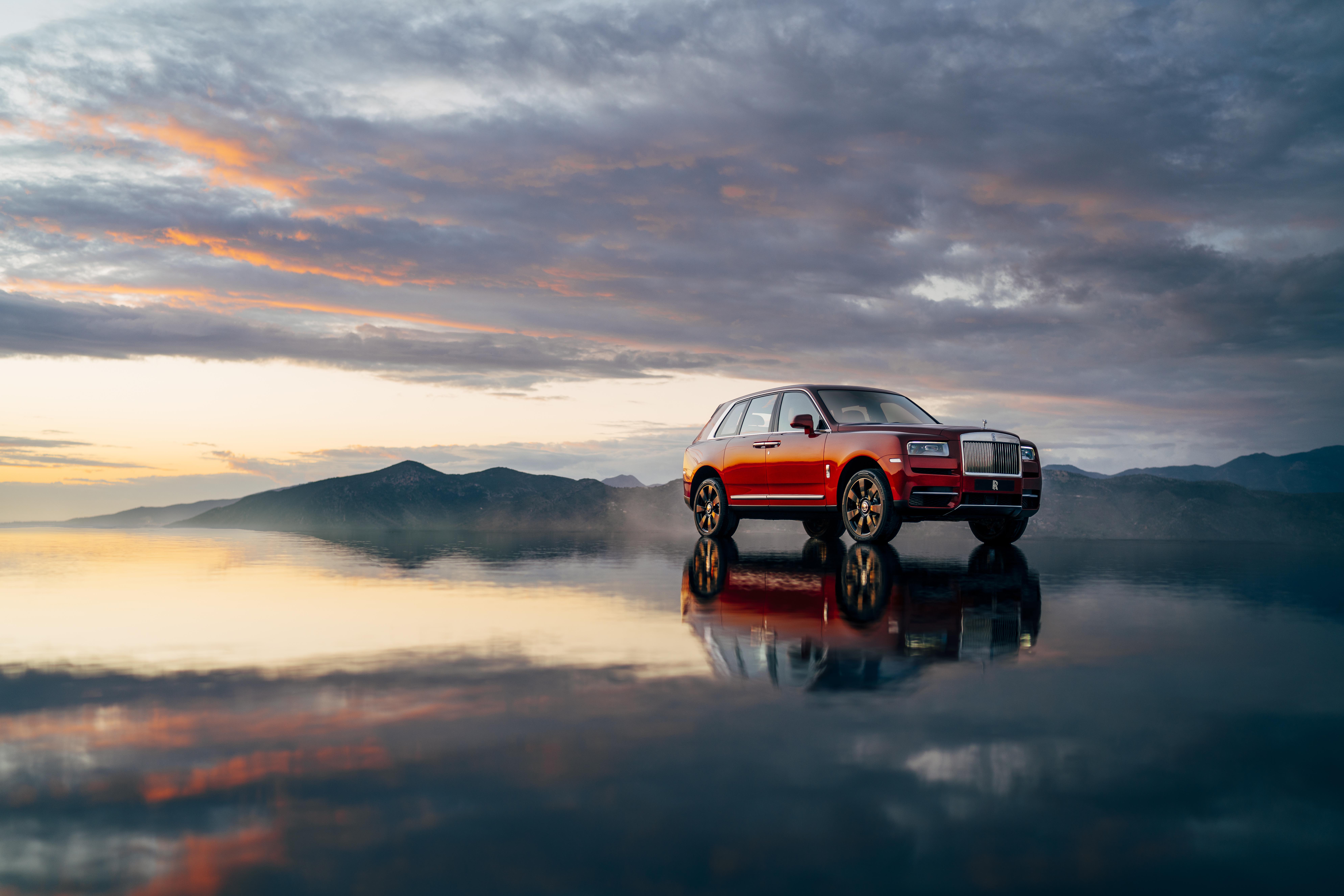Utilitário esportivo Rolls-Royce Cullinan na cor laranja em um chão molhado com reflexo do carro e do céu e montanhas ao fundo no entardecer 