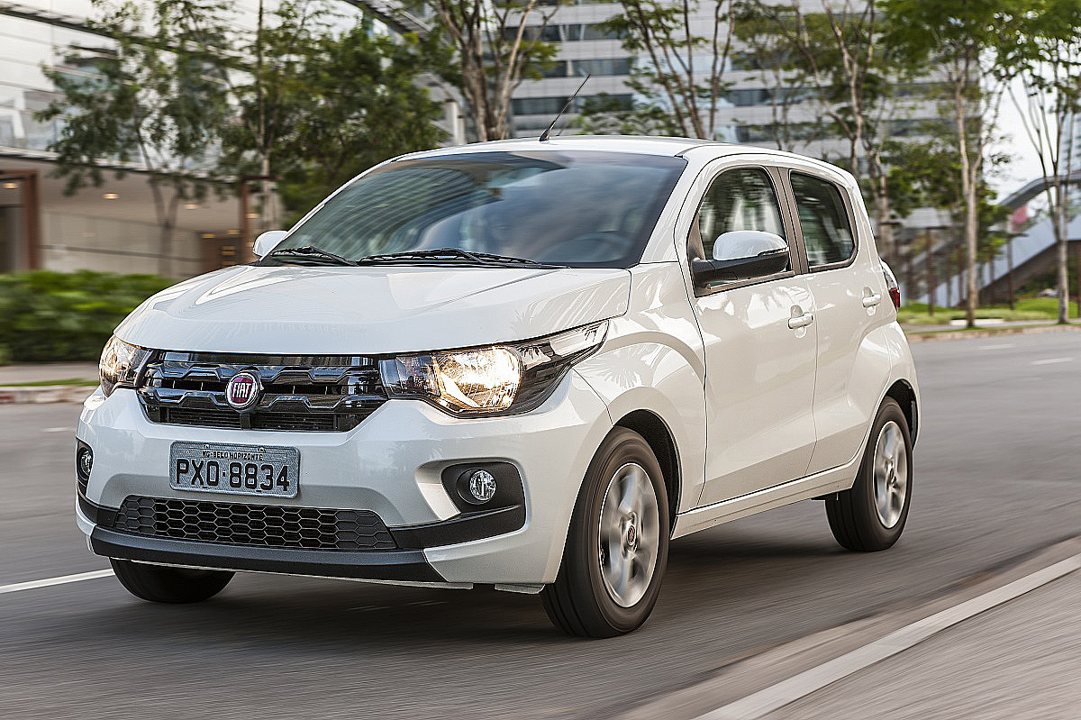 Subcompacto Fiat Mobi branco, quatro portas, com farois acesos na rua durante o dia