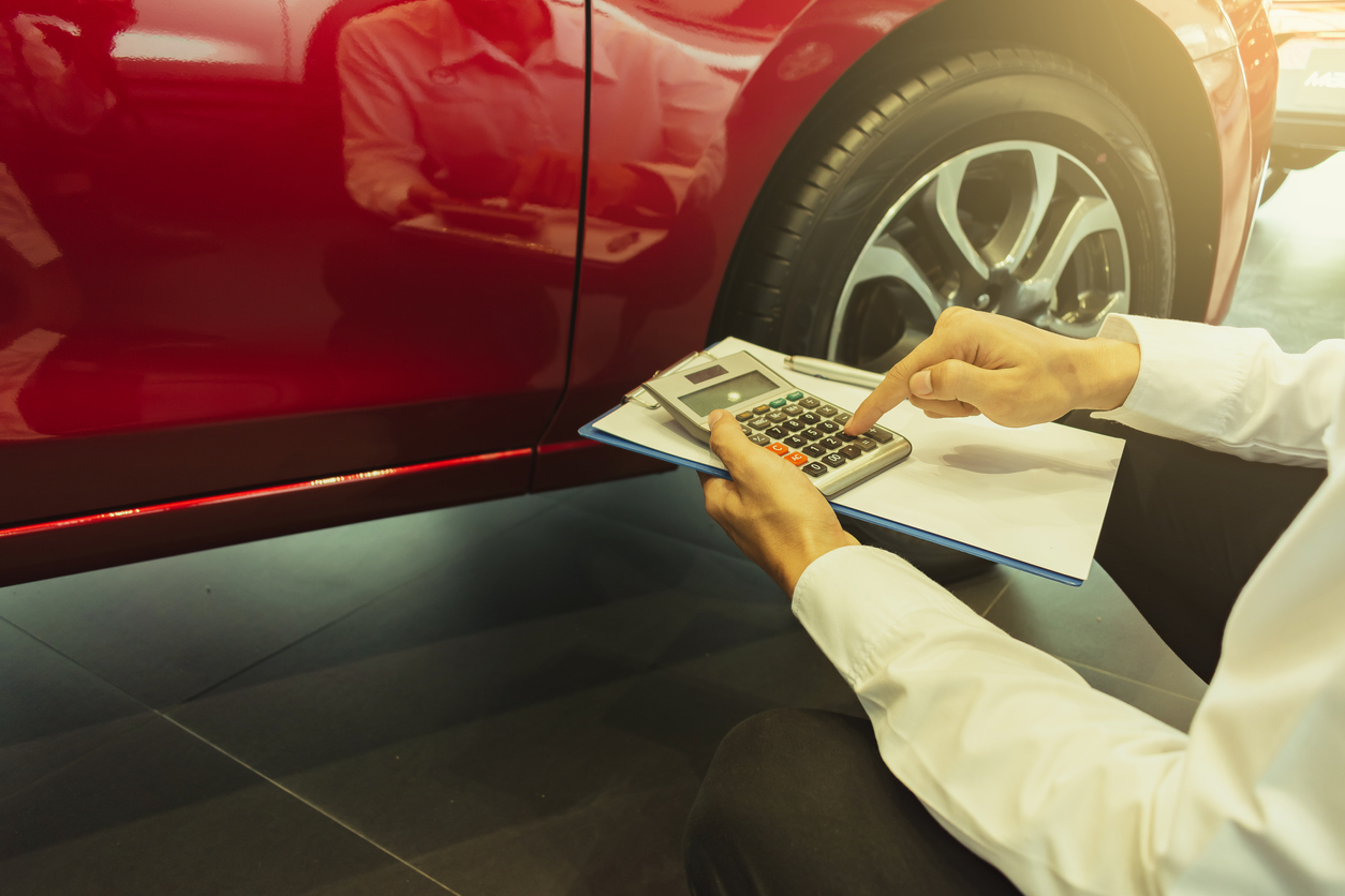 Homem segura uma calculadora e um bloco de notas em frente a um carro vermelho