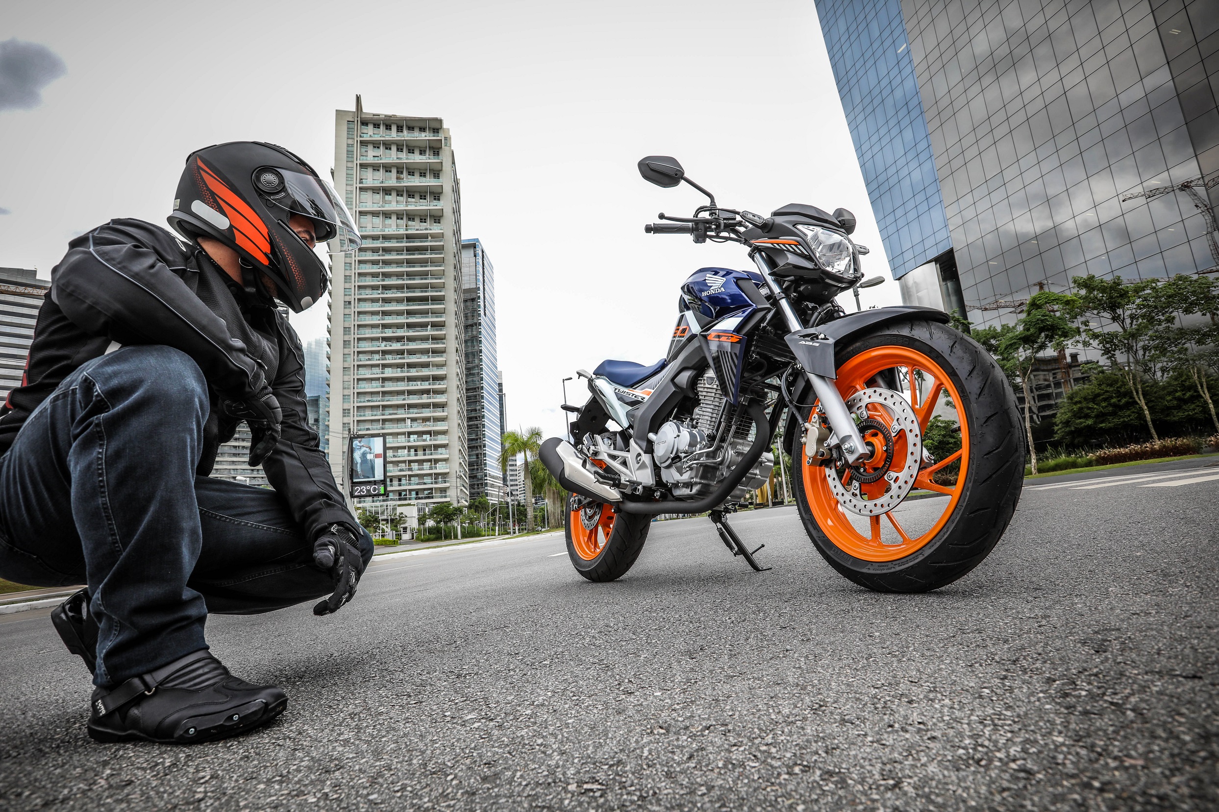 Homem de capacete observa a Honda CB250 Twister Special Edition fotografada de baixo com as rodas laranjas e tanque azul