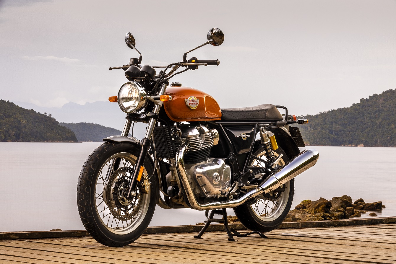 Royal Enfield Interceptor 650 estática de frente com seu desenho clássico, guidom reto, espelhos com hastes longas, tanque laranja tendo ao fundo um lago