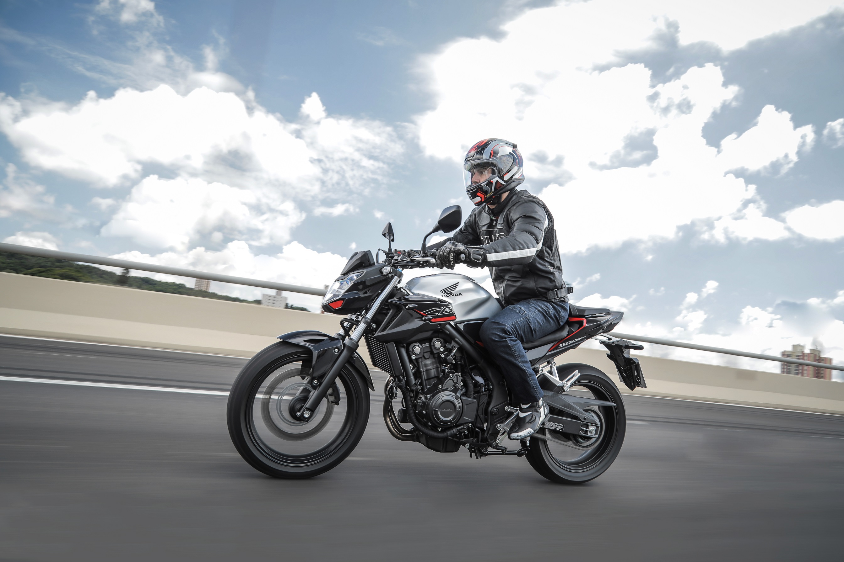 Honda CB 500F sendo pilotada em movimento com tanque prata e sobre viaduto