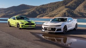 Ford Mustang verde ao lado de um Chevrolet Camaro branco tendo ao fundo um lago cercado de montanhas