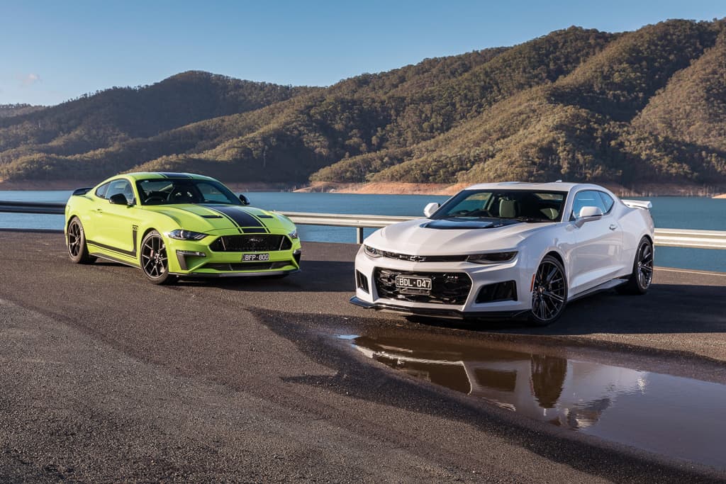 Ford Mustang verde ao lado de um Chevrolet Camaro branco tendo ao fundo um lago cercado de montanhas