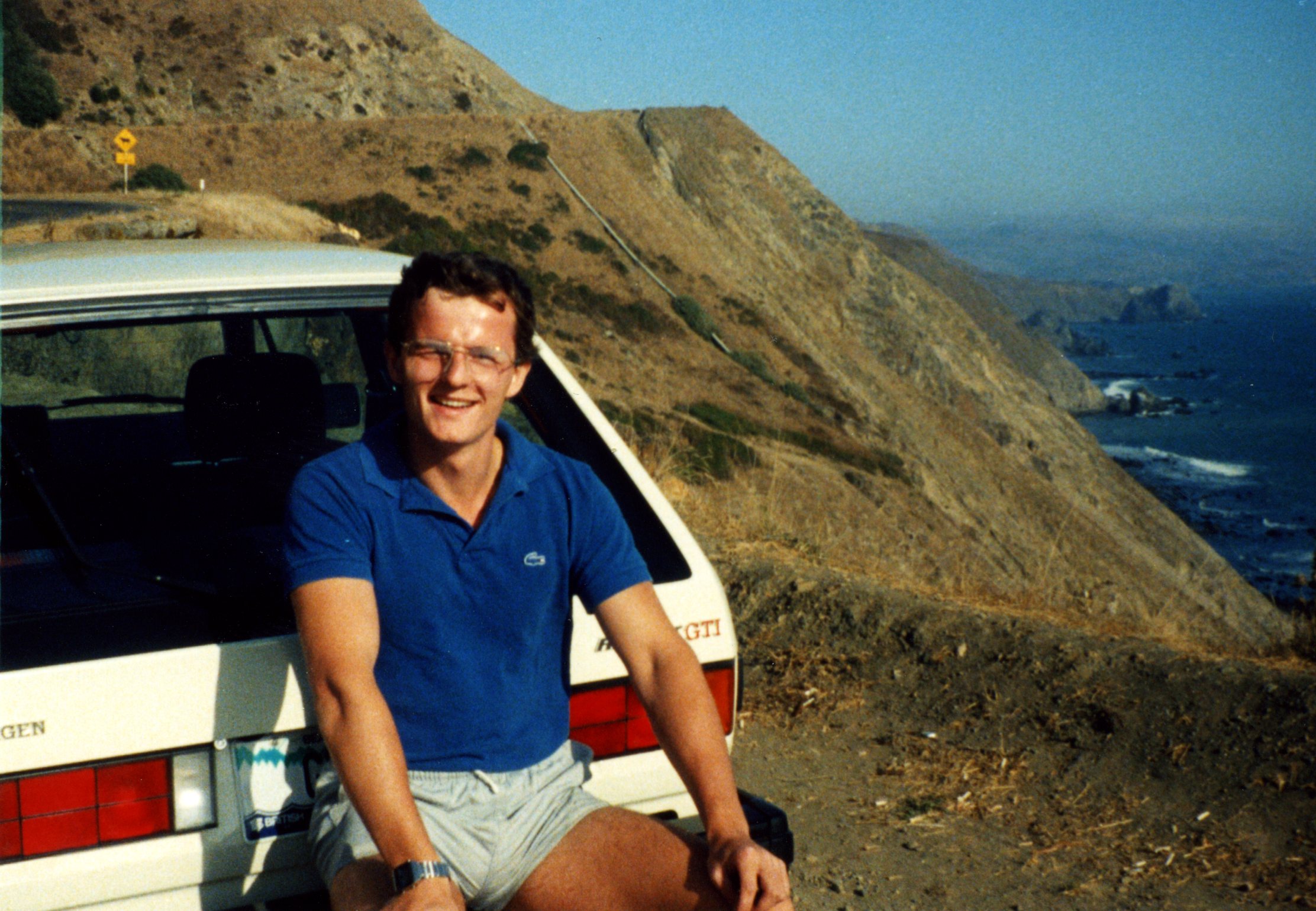 Derek Pratt sentado no para-choque traseiro do seu Volkswagen Golf Gti original branco e ao fundo montanhas e um vale