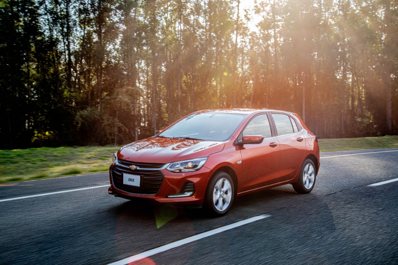 Chevrolet Onix vermelho em movimento na estrada com sol baixo atrás de uma plantação de eucalliptos