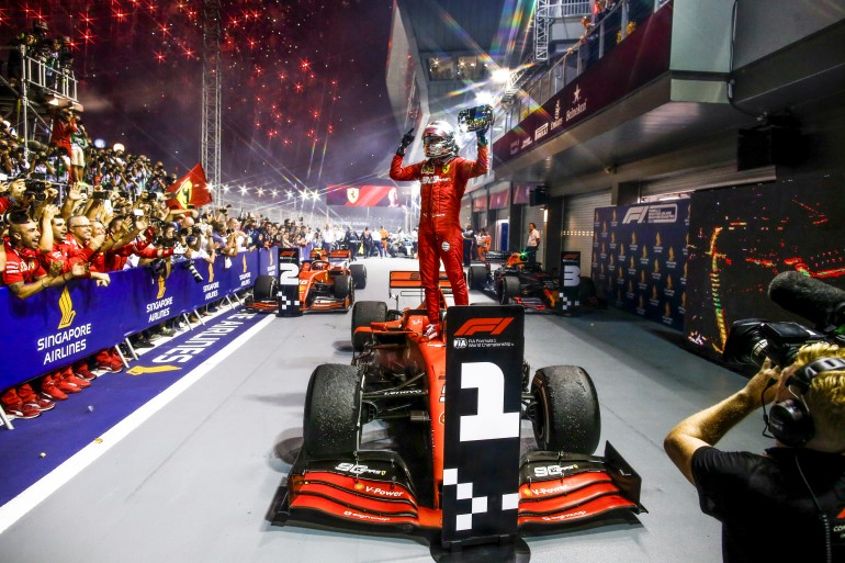 Vettel sobe no carro da Ferrari, nos boxes do GP de Singapura 2019 2, de macacão vermelho, capacete e volante na mão para comemorar a vitória