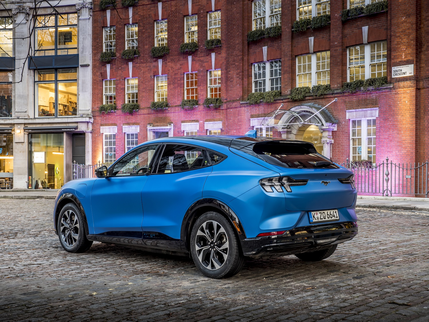 Ford Mustang Mach-E azul de traseira estacionado em uma rua na frente de um prédio clássico com fachada vinho