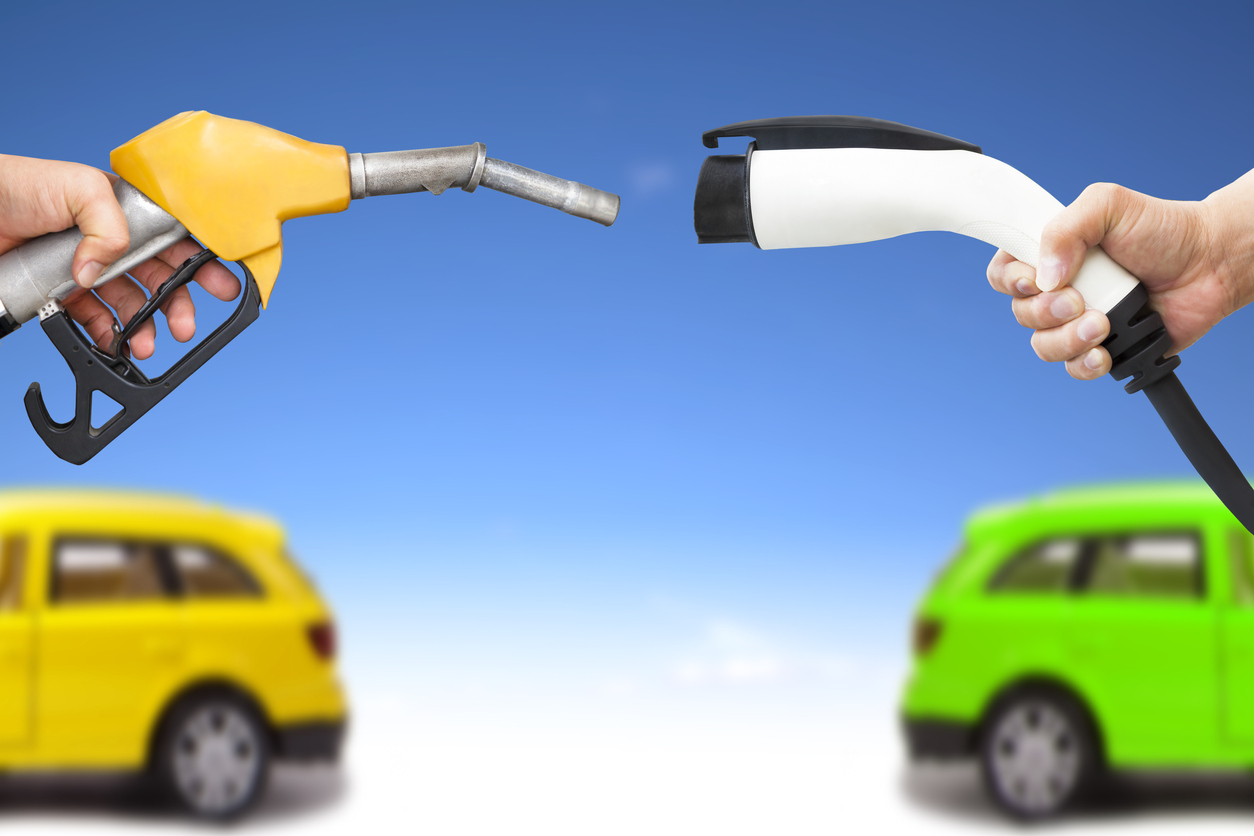 Monatem mostra um bico de bomba de gasolina amarelo de frente para um soquete de carregamento de carro elétrico branco ao fundo a traseira de um carro amarelo e a tarseira de um carro verde, ambos desfocados