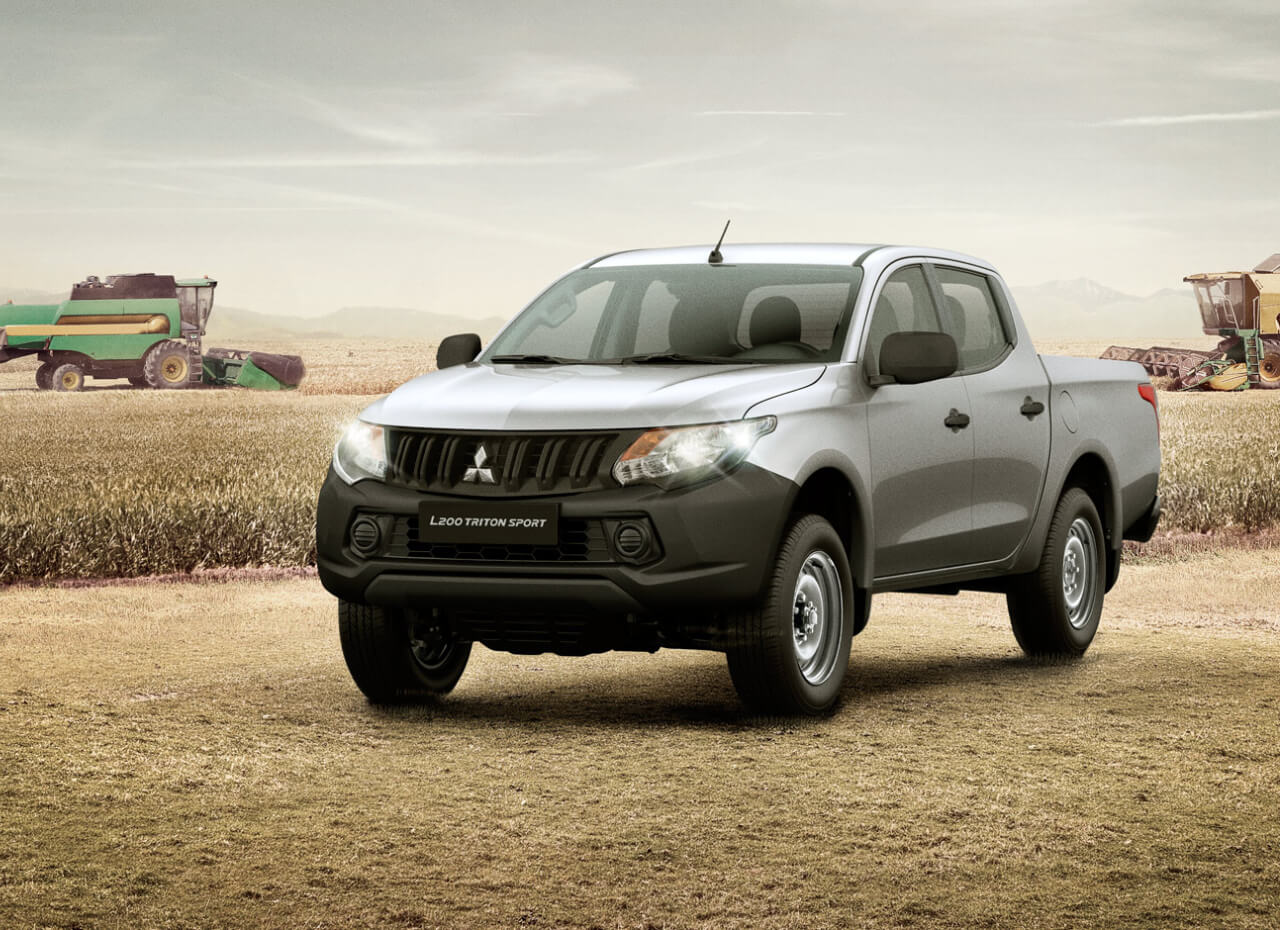 Mitsubishi L200 Triton prata de frente em cenário de plantação tendo ao fundo um trator e uma semeadeira