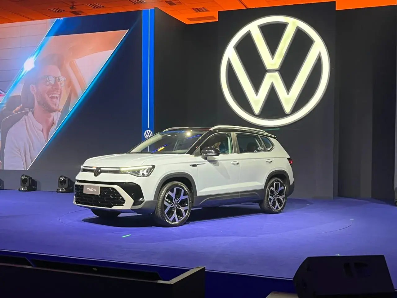 Volkswagen Taos 2026 na cor branca com teto preto em posição 3/4 de frente. Logo da Volkswagen se destaca no funto