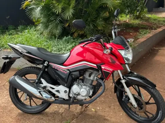 Imagem de uma Honda CG 160 Titan vermelha estacionada ao ar livre, próxima a plantas verdes, com preço até 18 mil reais.