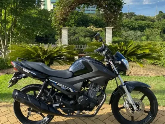 Imagem de uma Yamaha Factor 150ED preta estacionada em frente a um jardim com árvores verdes e céu azul ao fundo, perfeita para quem busca economia de combustível e desempenho em até 18 mil reais.
