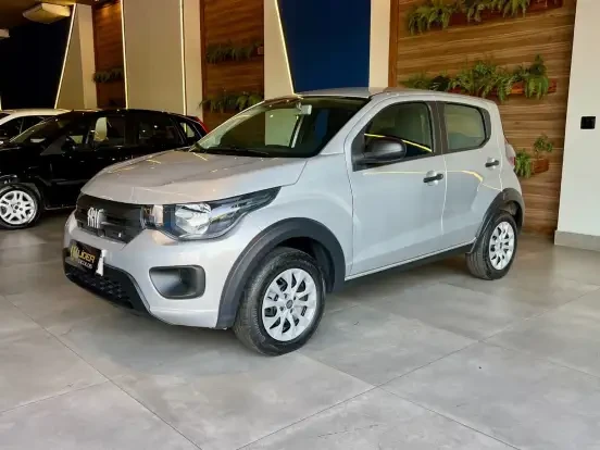 Fiat Mobi branco em showroom com decoração moderna e veículos ao fundo, destacando-se por seu design compacto e eficiente para uso urbano.