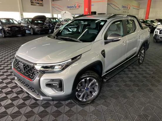 Imagem de uma Fiat Strada prata em showroom, destacando seu design moderno e compacto ideal para trabalho e lazer, em ambiente com outros veículos ao fundo.