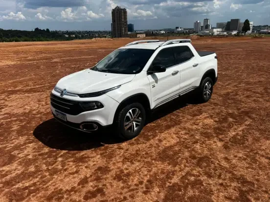Imagem de uma Fiat Toro branca estacionada em um terreno árido com vista para a cidade ao fundo, destaque para seu design robusto e moderno.